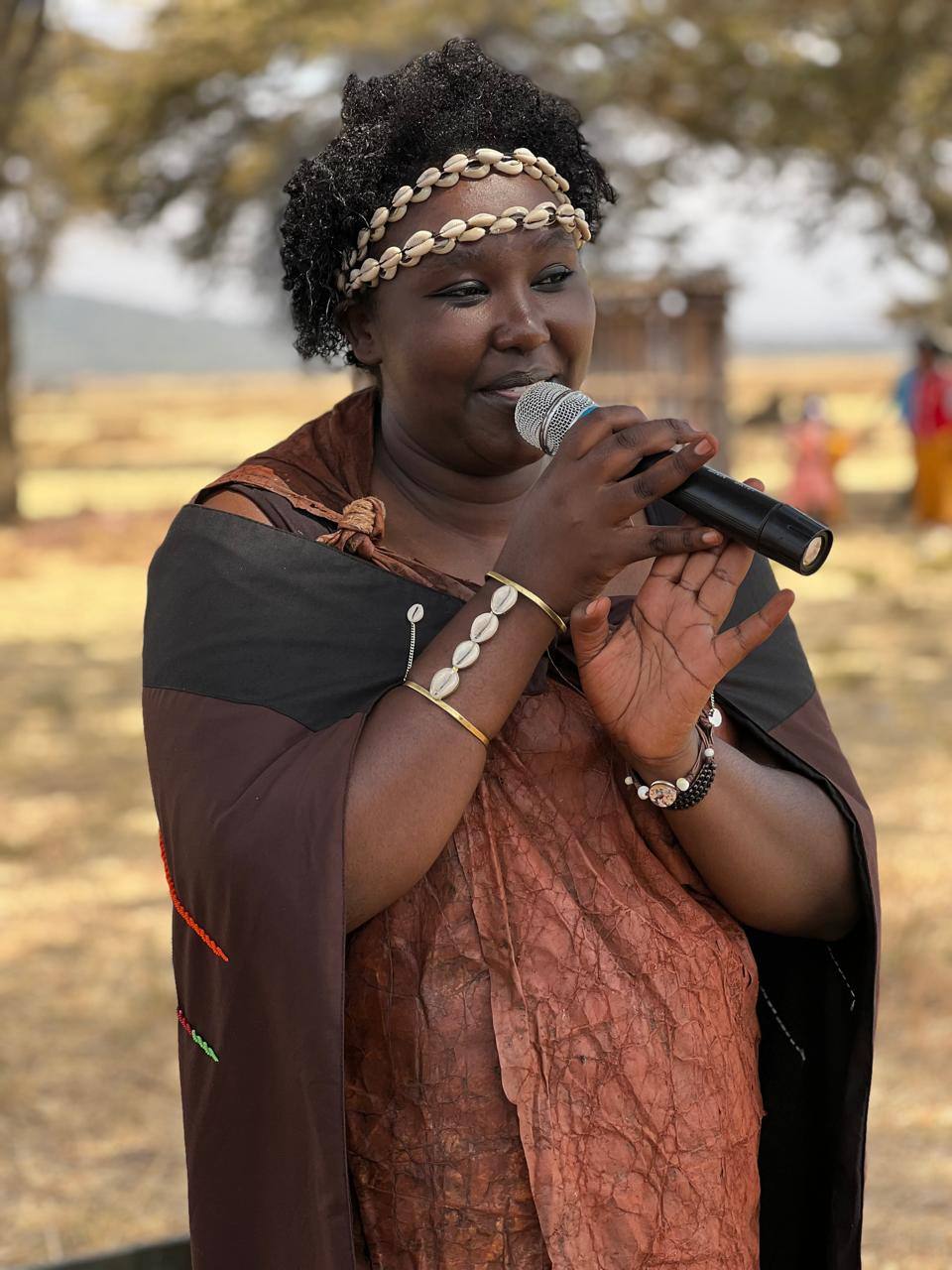 Juliana in a cowrie-shell headband and traditional hide dress speaking into a microphone at Dol Dol